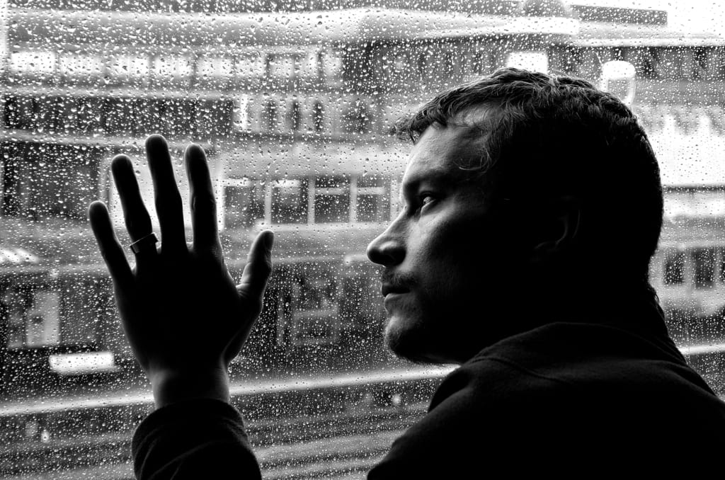 Hombre joven de costado viendo llover a traves de una ventana, refleccionando